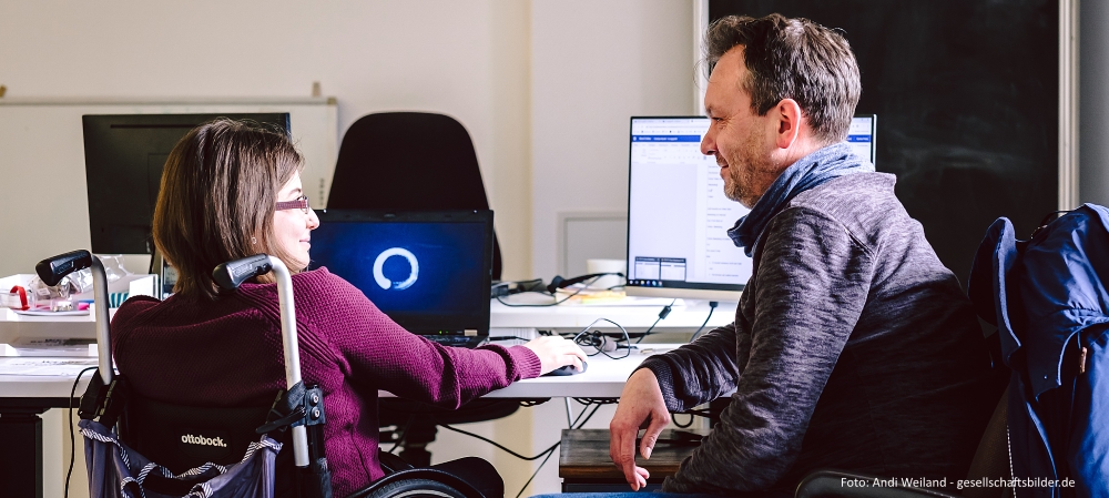 Zwei Menschen sitzen an einem Schreibtisch und unterhalten sich.